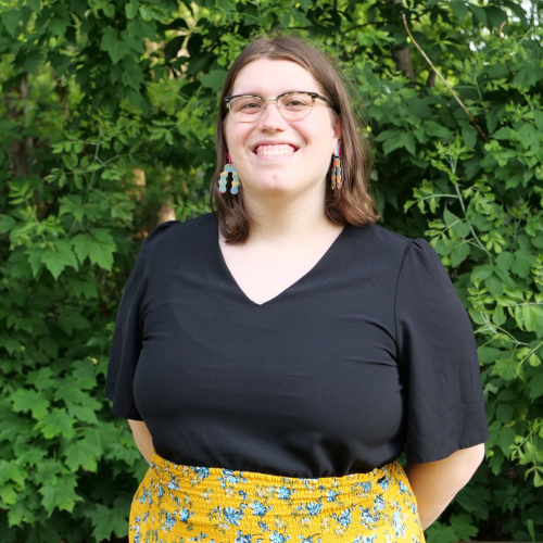 A photo of me wearing a yellow floral skirk and a flowy black top. The background is viney bush